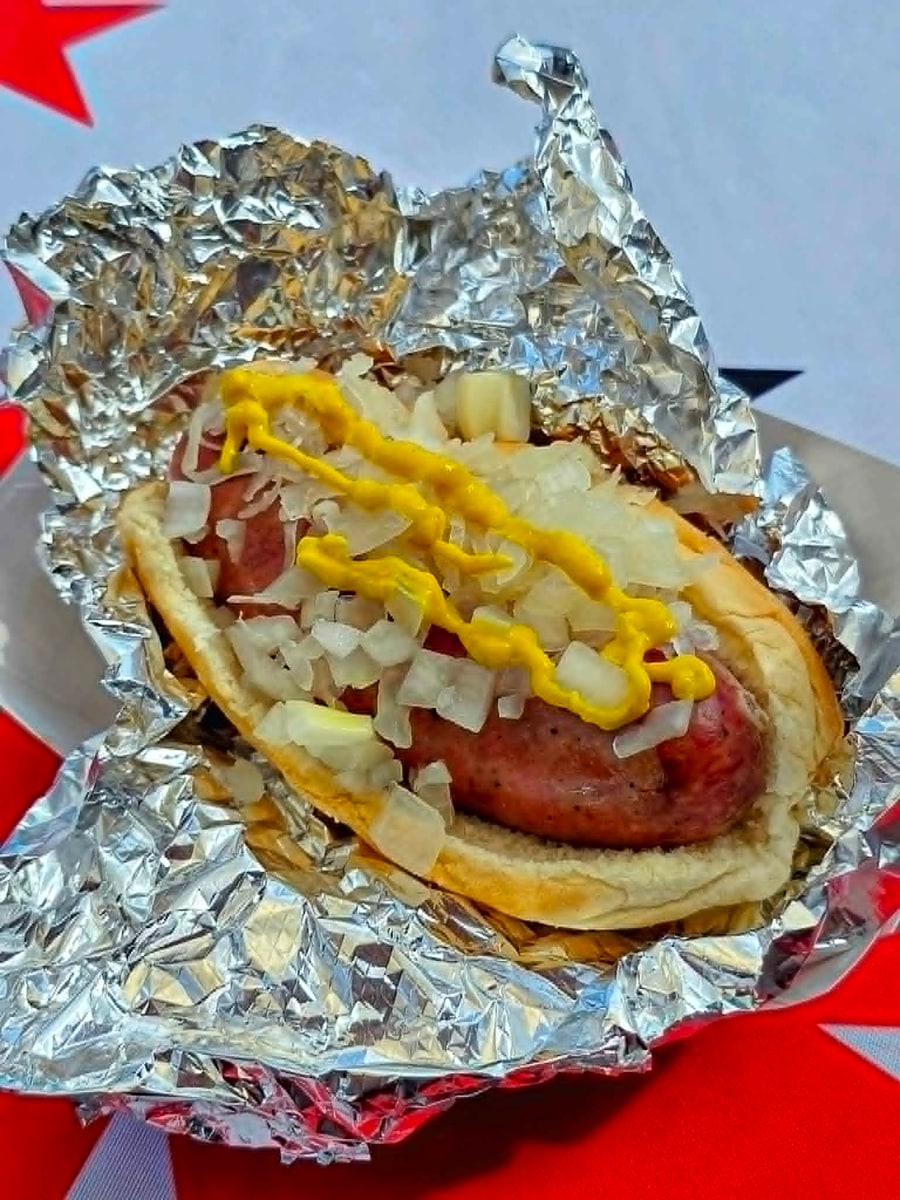 Brat supreme topped with melted cheese, diced onion, and mustard